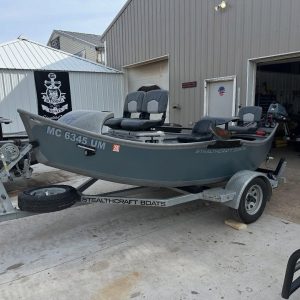 Gray Stealthcraft boat on a trailer outside a workshop, with two front seats and an outboard motor visible inside the hull area.