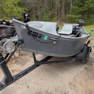 Gray fishing boat on a trailer in a muddy dirt lot, with a black outboard motor and forest in the background
