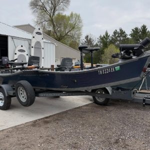 Blue StealthCraft fishing boat on a trailer in a gravel yard next to a metal workshop, with fishing gear and seats visible in the boat.