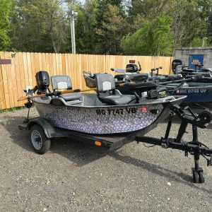 Several aluminum fishing boats on trailers parked in a lot beside a wooden fence and trees; outboard motors visible on the boats.