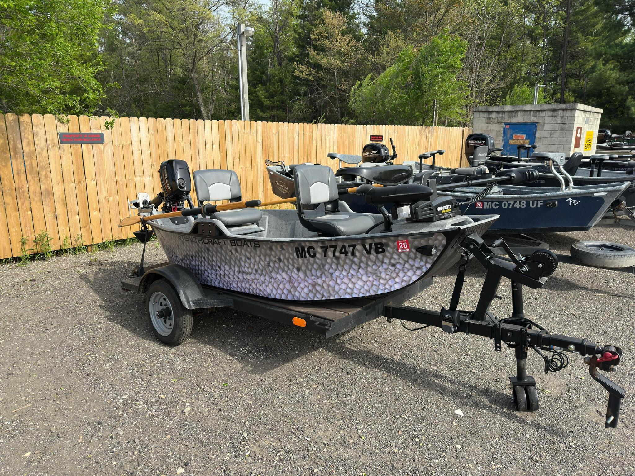 Several aluminum fishing boats on trailers parked in a lot beside a wooden fence and trees; outboard motors visible on the boats.