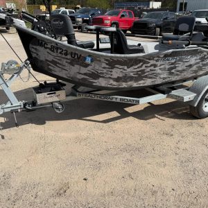 Camouflage-patterned fishing boat on a trailer in a dirt lot, with several pickup trucks parked in the background.