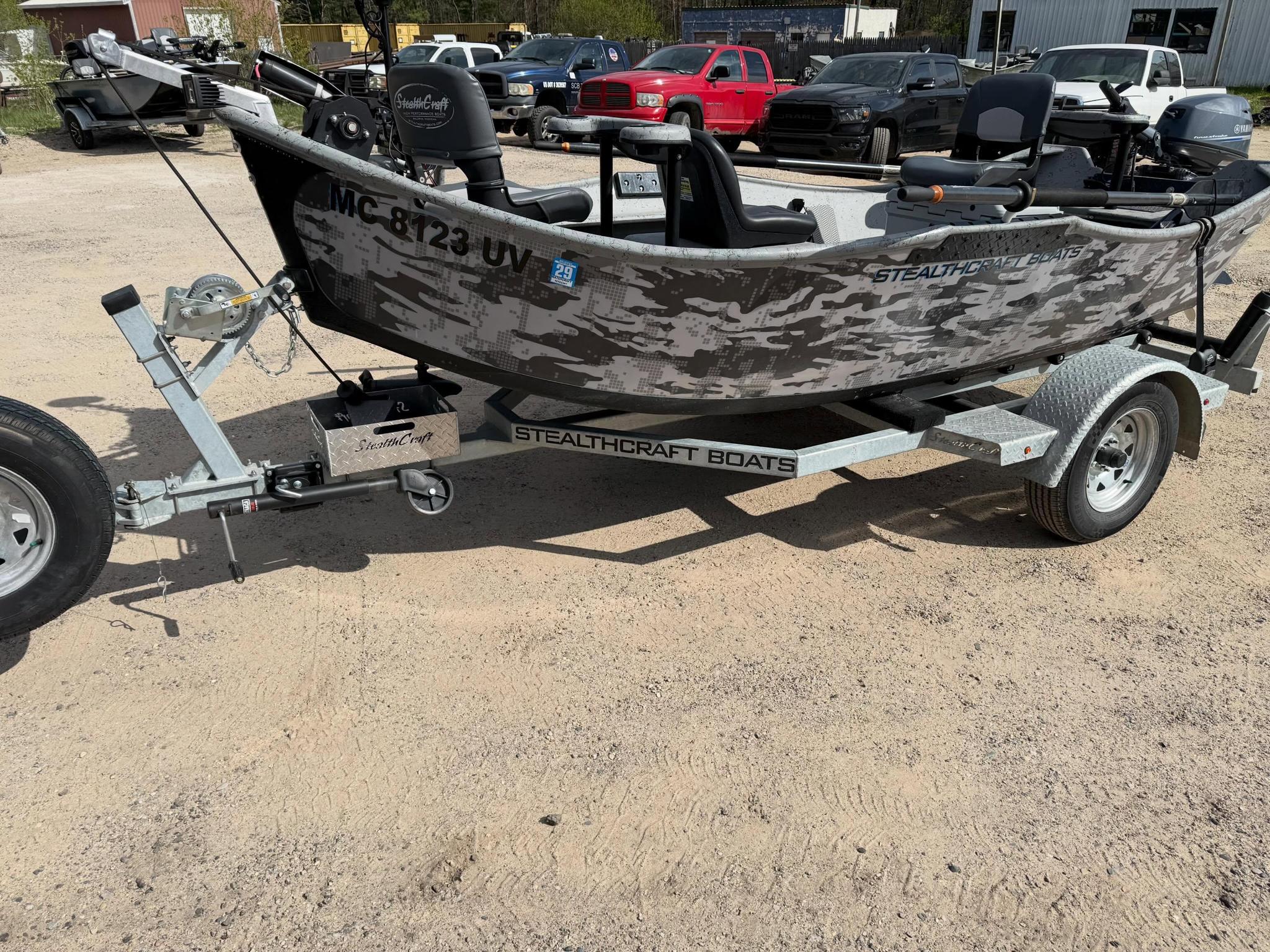 Camouflage-patterned fishing boat on a trailer in a dirt lot, with several pickup trucks parked in the background.
