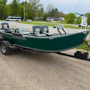 Green fishing boat on a black trailer in a gravel parking lot, with multiple grey seats and a trolling motor mounted at the bow.