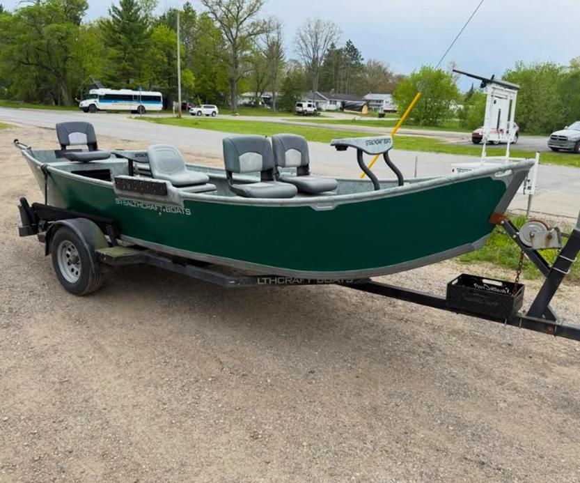 Green fishing boat on a black trailer in a gravel parking lot, with multiple grey seats and a trolling motor mounted at the bow.