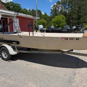 Beige boat on a trailer in a parking lot beside a red building; outboard motors visible on the transom and a reserved parking sign nearby.