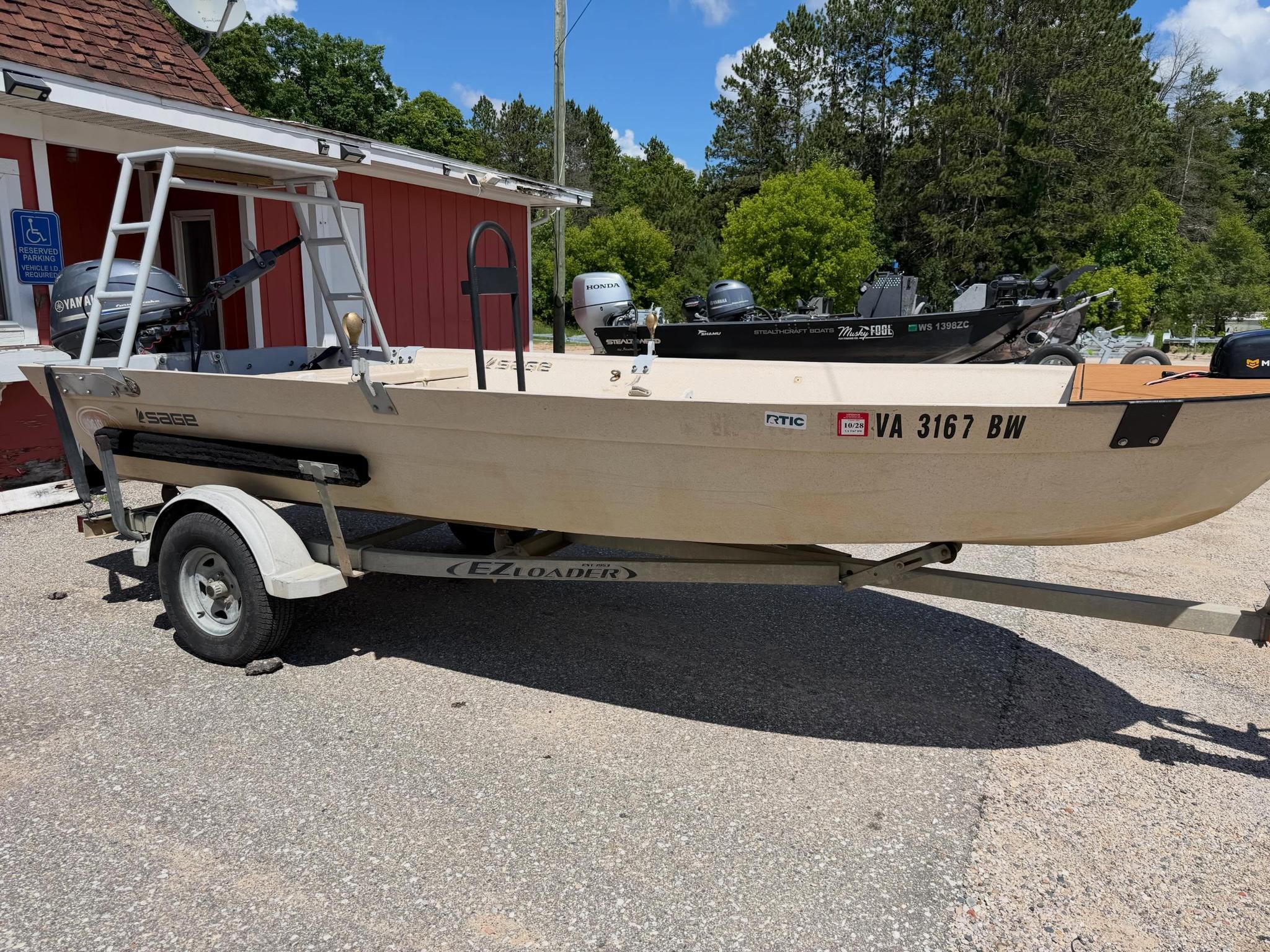 Beige boat on a trailer in a parking lot beside a red building; outboard motors visible on the transom and a reserved parking sign nearby.