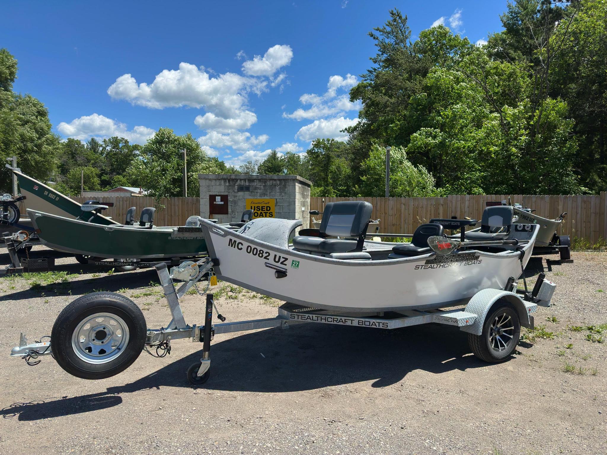 Row of boats on trailers parked in a boatyard with a wooden fence, under a bright blue sky with scattered clouds. Ready for storage or transport.