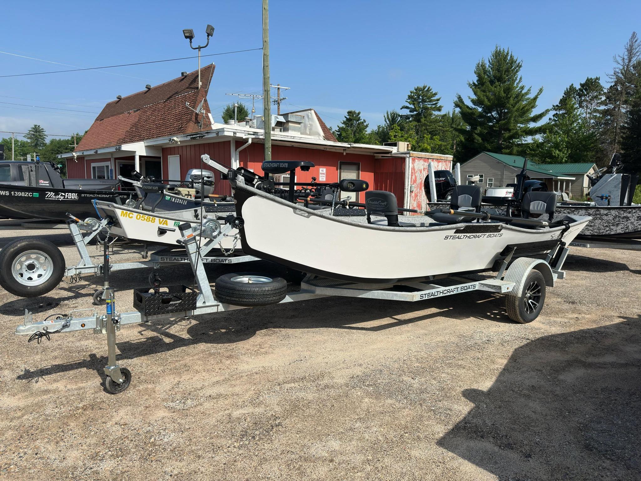 Boats on trailers in a boatyard, with a red shed and green trees in the background under a clear blue sky.