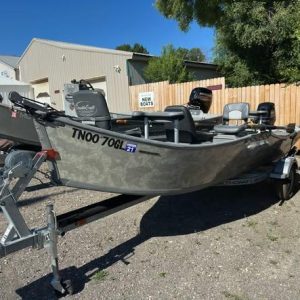 Aluminum fishing boat on a trailer in a gravel lot, with seats and an outboard motor, parked near a wooden fence and small buildings; a sign reading 'New Boats' in the background.