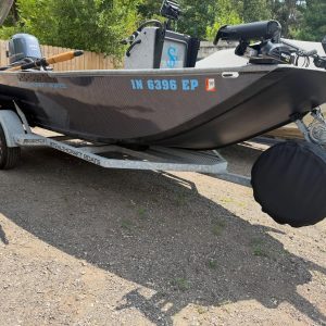 Black and blue fishing boat on a trailer in a gravel lot, with a cover over the outboard motor and gear mounted at the bow and stern.