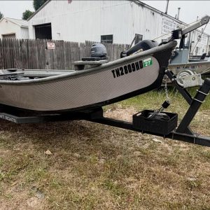 Aluminum fishing boat on a black trailer in a gravel yard beside a wooden fence and a white metal building with outboard motors attached to the stern.