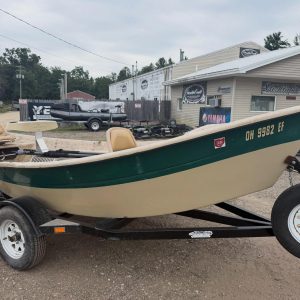 Green and beige speedboat on a trailer in a boatyard with a building and signs in the background.