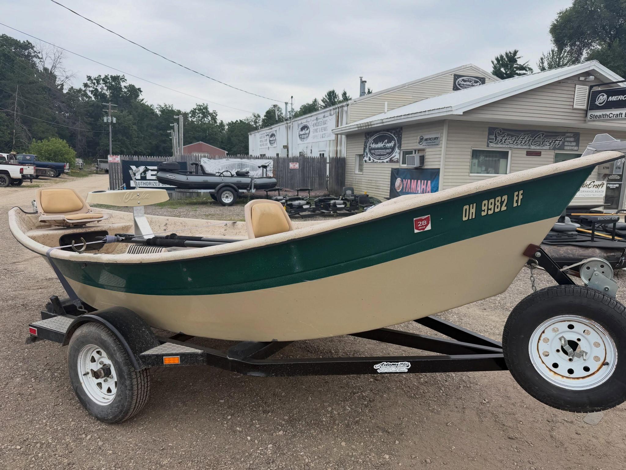 Green and beige speedboat on a trailer in a boatyard with a building and signs in the background.