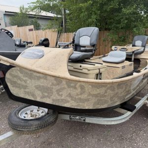 Camouflage boat on a trailer in a gravel lot, with gray cockpit seats and a Mercury motor nearby behind a wooden fence.