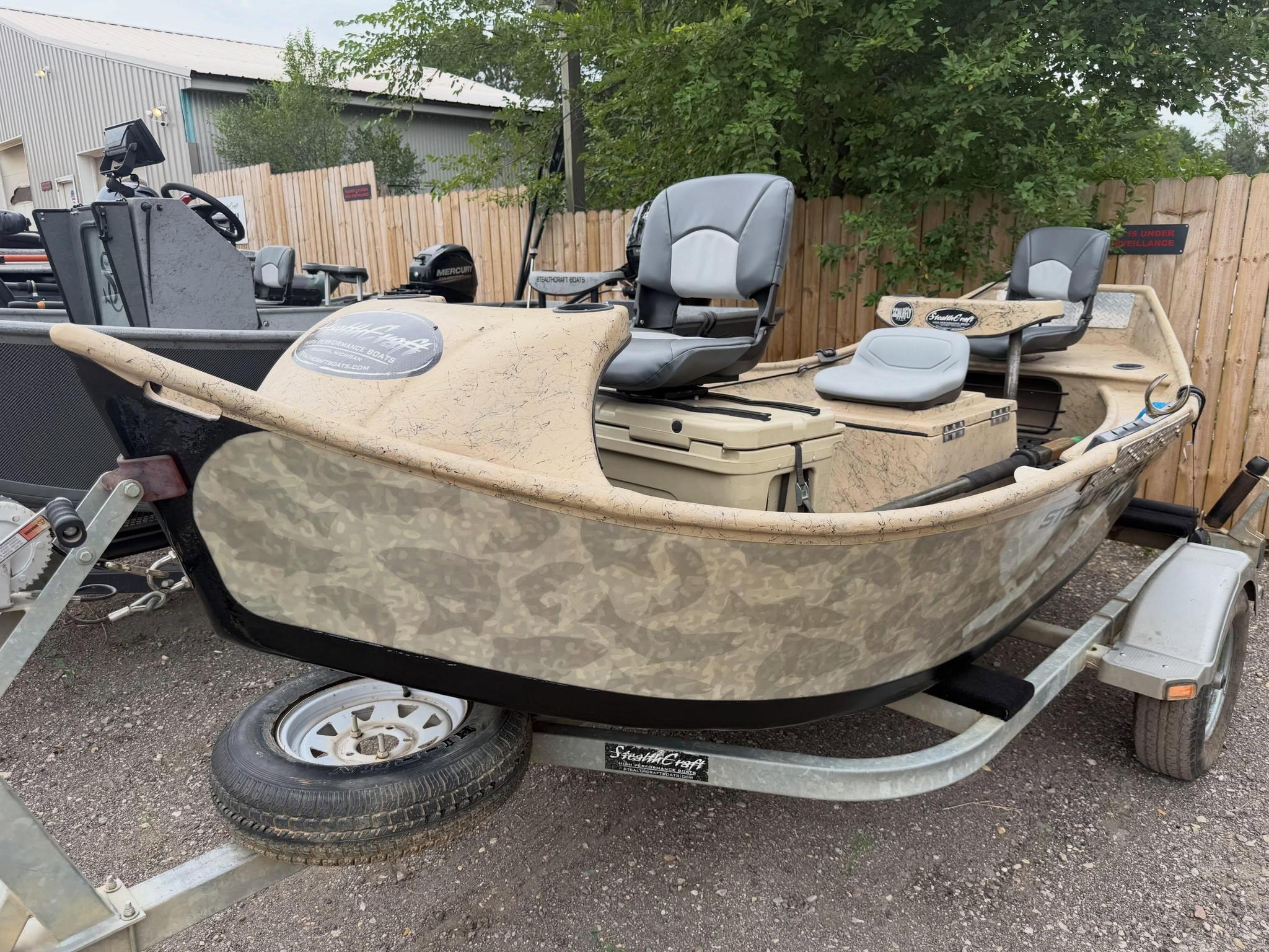 Camouflage boat on a trailer in a gravel lot, with gray cockpit seats and a Mercury motor nearby behind a wooden fence.