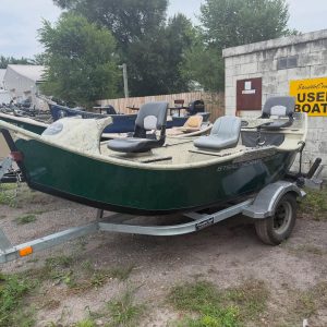 Boats on a trailer in a boatyard with a yellow 'USED BOATS' sign on a concrete wall behind them.