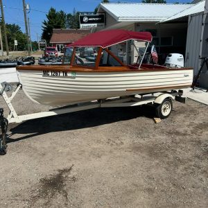 White wooden speedboat on a trailer outside a shop, red canopy and polished wooden trim, registration MC 1267 TK visible on the hull.
