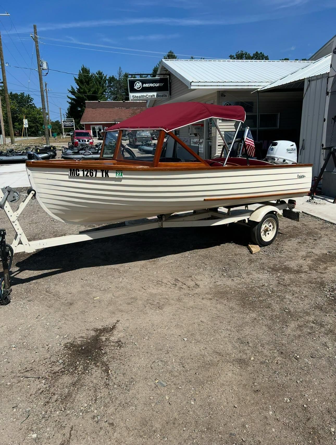 White wooden speedboat on a trailer outside a shop, red canopy and polished wooden trim, registration MC 1267 TK visible on the hull.